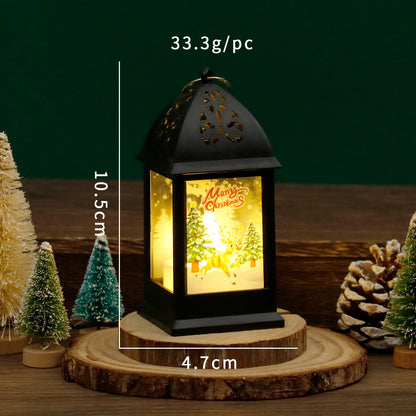 Decorative lantern with Christmas design on a wooden base, surrounded by small trees and pinecones.