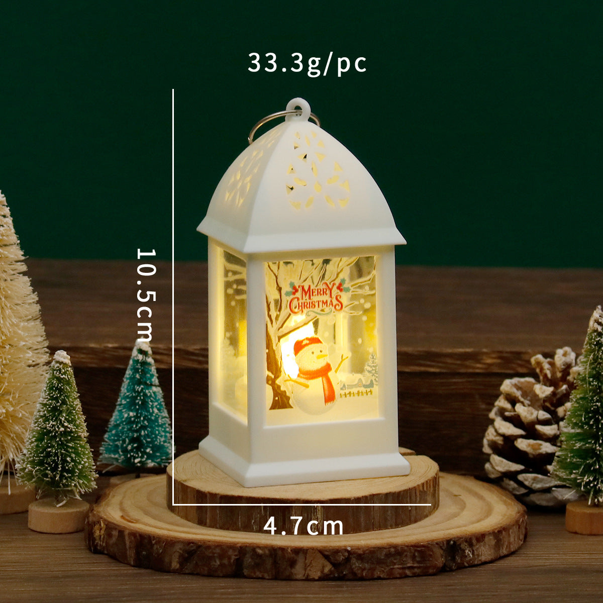 White lantern with Christmas design on a wooden base, surrounded by small trees and pine cones.