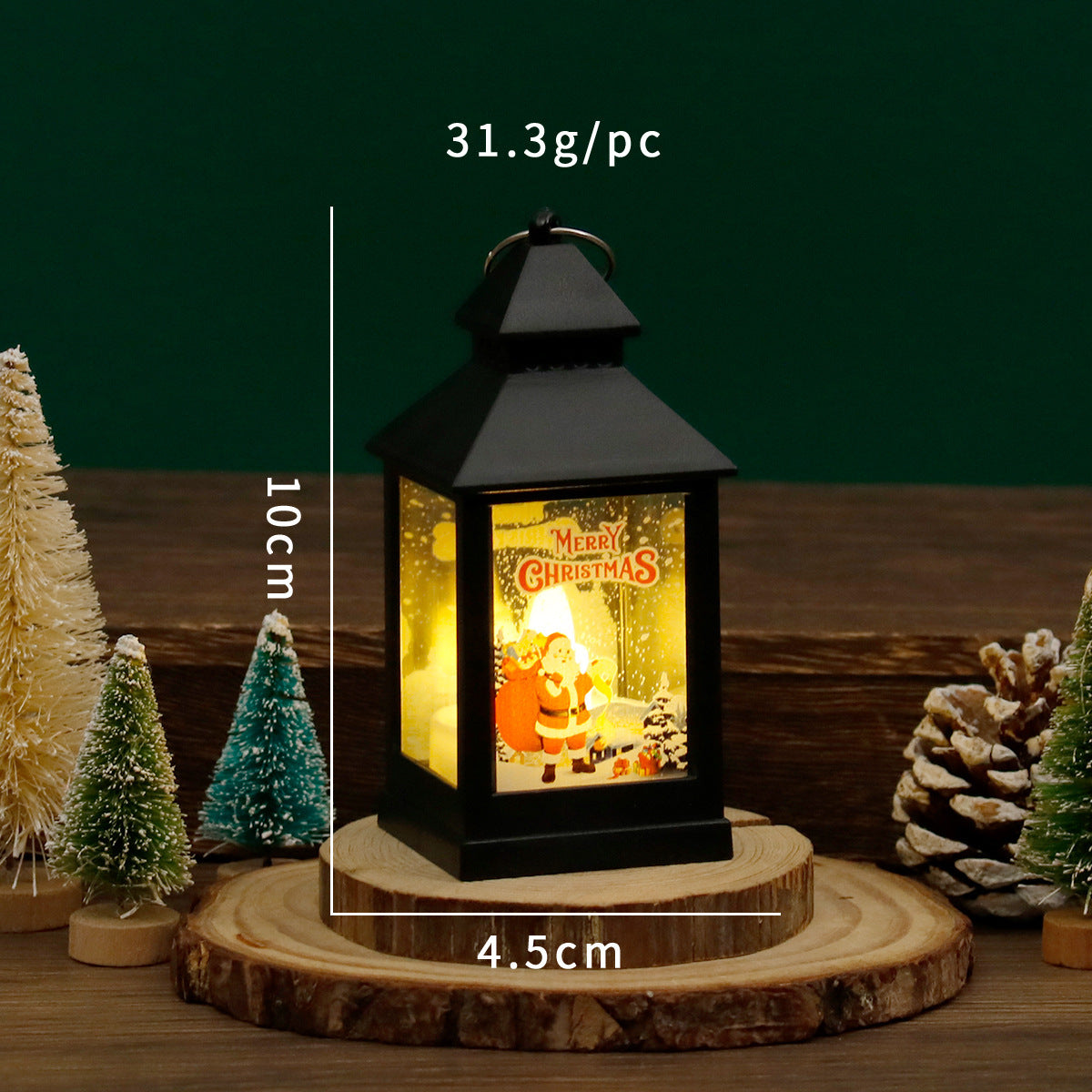 Decorative lantern with Christmas design on a wooden base, surrounded by small trees and pine cones.
