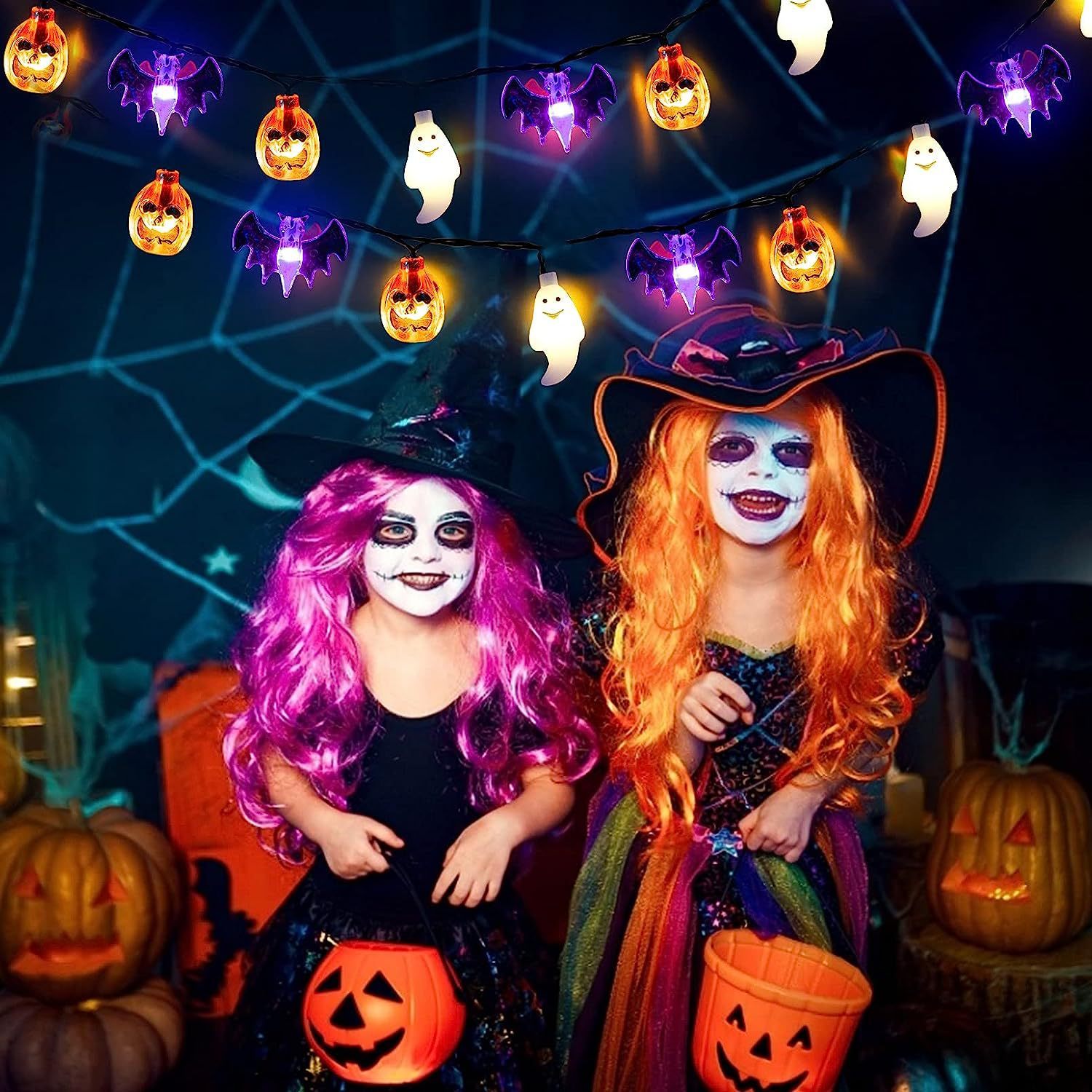 Two children in Halloween costumes with colorful wigs and pumpkin buckets, surrounded by Halloween lights and decorations.